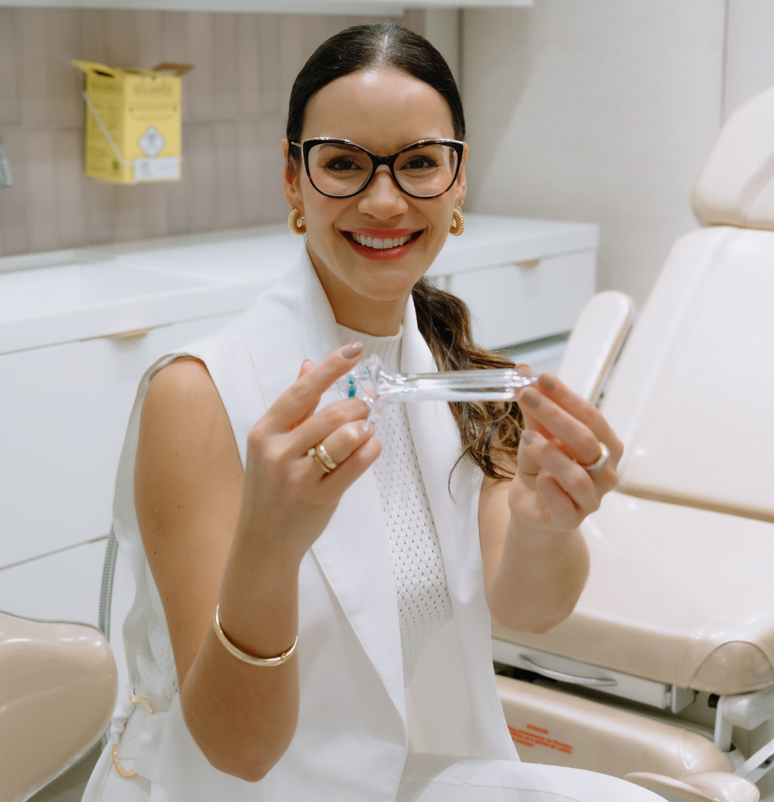 Dra. Marianne Costa, ginecologista, sorrindo e segurando um espéculo ginecológico em consultório.